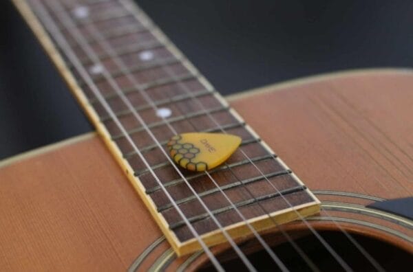 A Customized Guitar Pick Personalized in yellow rests on the fretboard of an acoustic guitar.