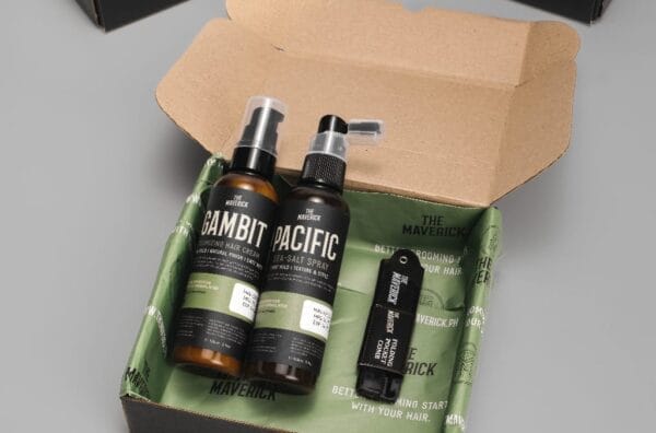 An open box containing The Maverick Gift Bundle 2, which includes Gambit face wash, Pacific sea salt spray, and a solid cologne called The Maverick. Boxes are visible in the background.