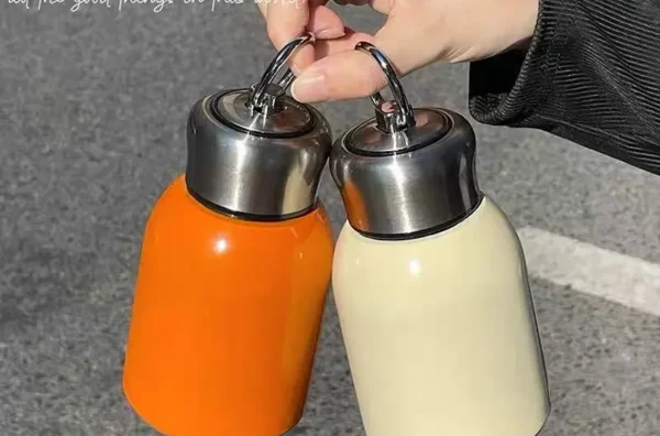 A hand is holding two Mini Couple Cup Tumblers with metal caps, one in orange and the other in cream-colored, against a gray background with a quote in cursive above.