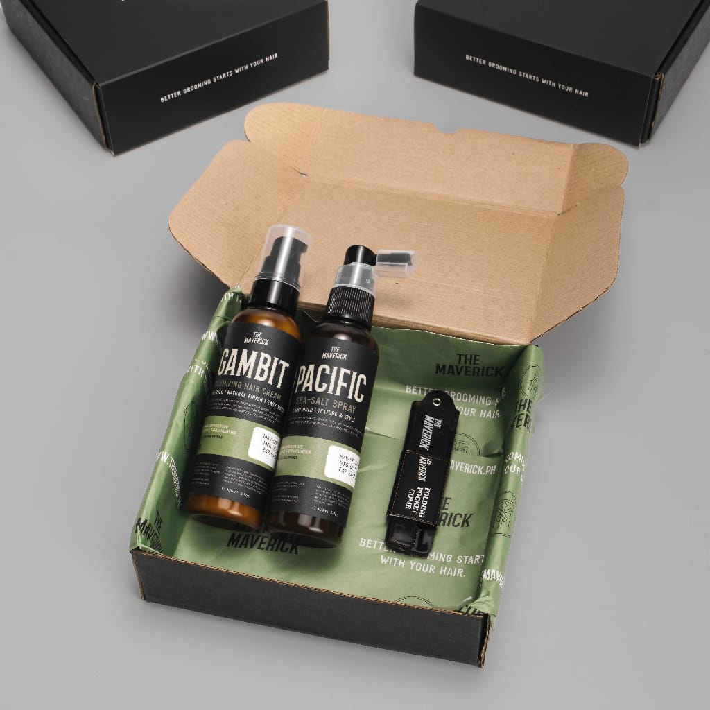 An open box containing The Maverick Gift Bundle 2, which includes Gambit face wash, Pacific sea salt spray, and a solid cologne called The Maverick. Boxes are visible in the background.