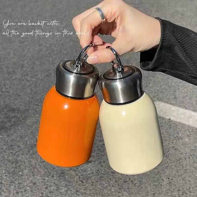 A hand is holding two Mini Couple Cup Tumblers with metal caps, one in orange and the other in cream-colored, against a gray background with a quote in cursive above.