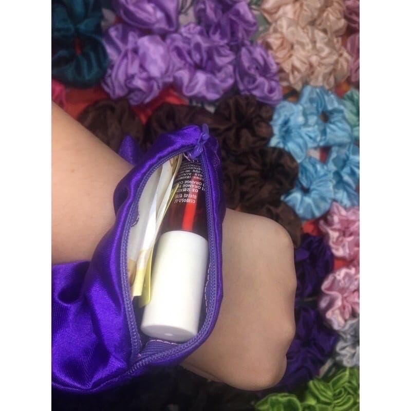 A silk Scrunchie With Pocket Zipper in purple velvet, featuring a hidden pocket that stores a small cylindrical item and a card, with an array of various colored scrunchies in the background.