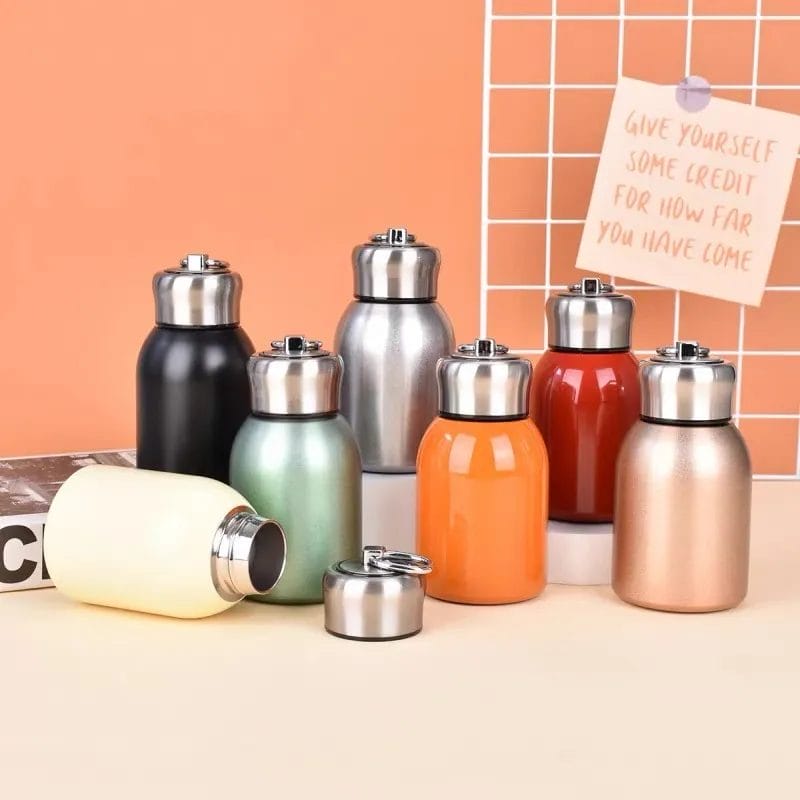 A collection of eight insulated stainless steel bottles in various colors are displayed on a flat surface, accompanied by a pair of Mini Couple Cup Portable Outdoor Tumblers and a wire grid holding a motivational note in the background.