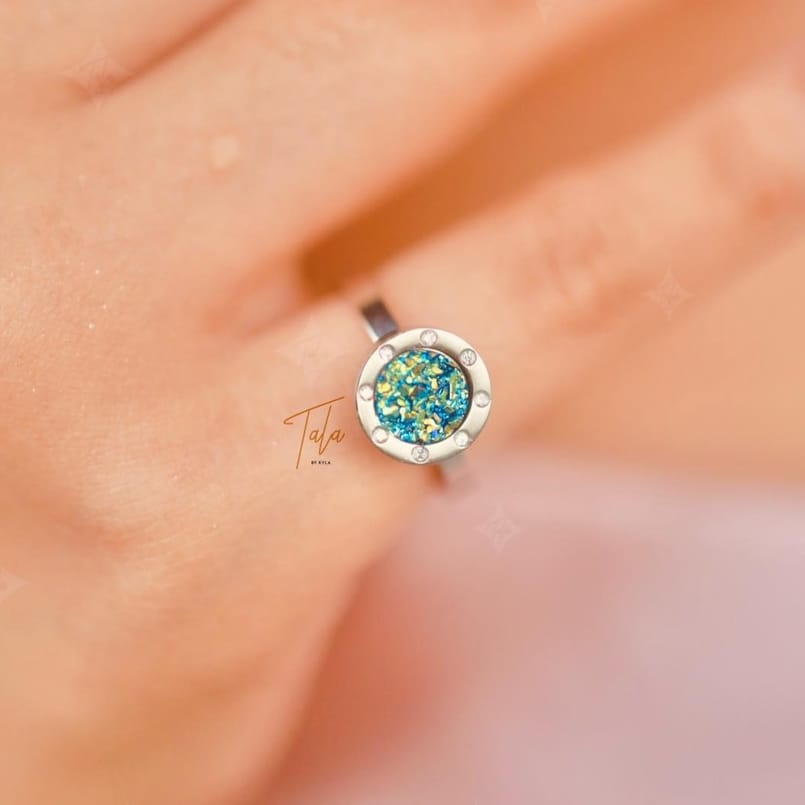 A woman's hand is holding a Tala by Kyla My Universe Fidget Ring with a blue stone.
