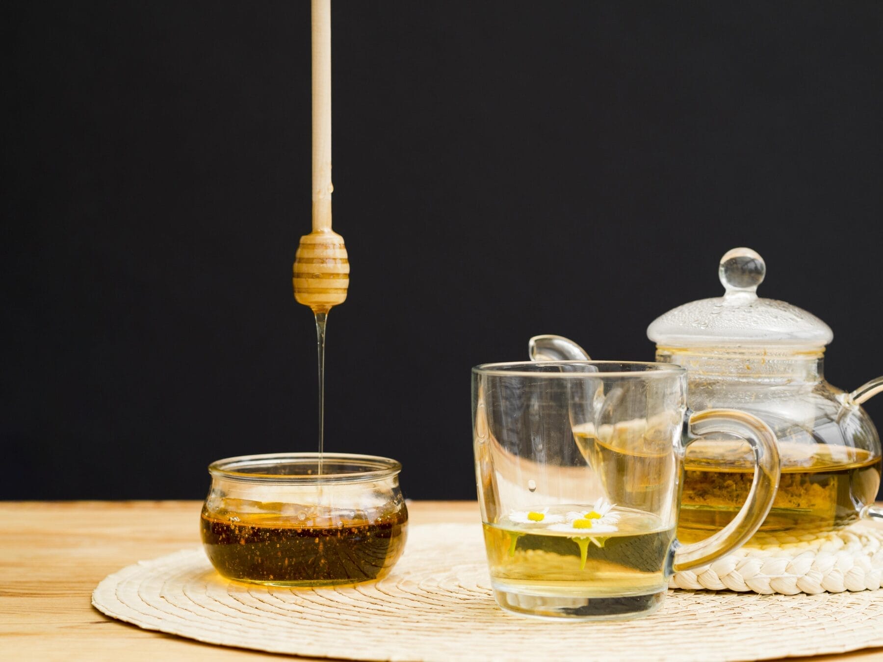 Honey drips from a dipper into a small jar, beside a glass cup of tea with chamomile flowers, and a glass teapot on a woven placemat against a dark background. honeymoon tea