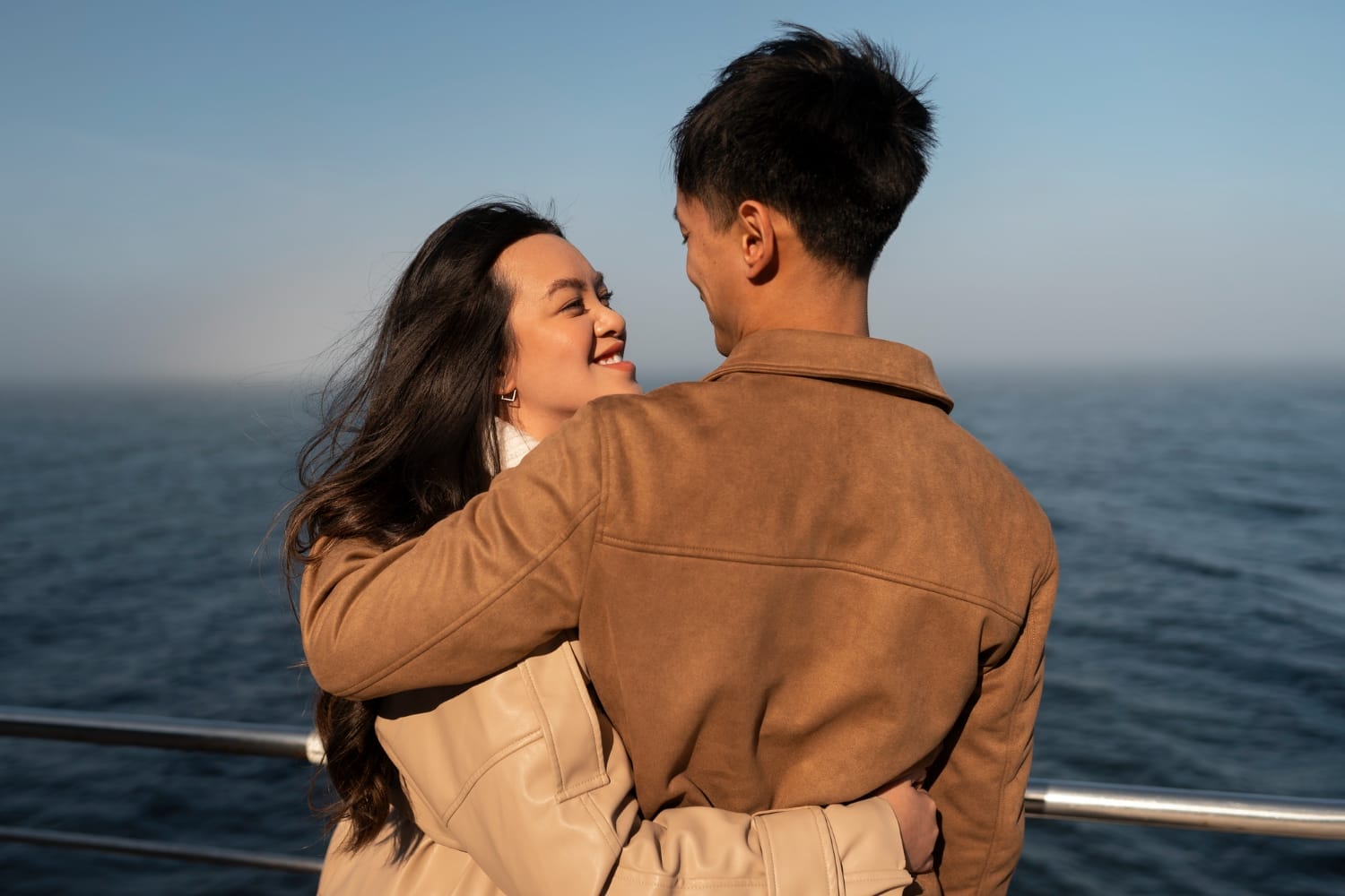 Infatuated, a couple embraces on a pier with the ocean sparkling behind them on a clear day.